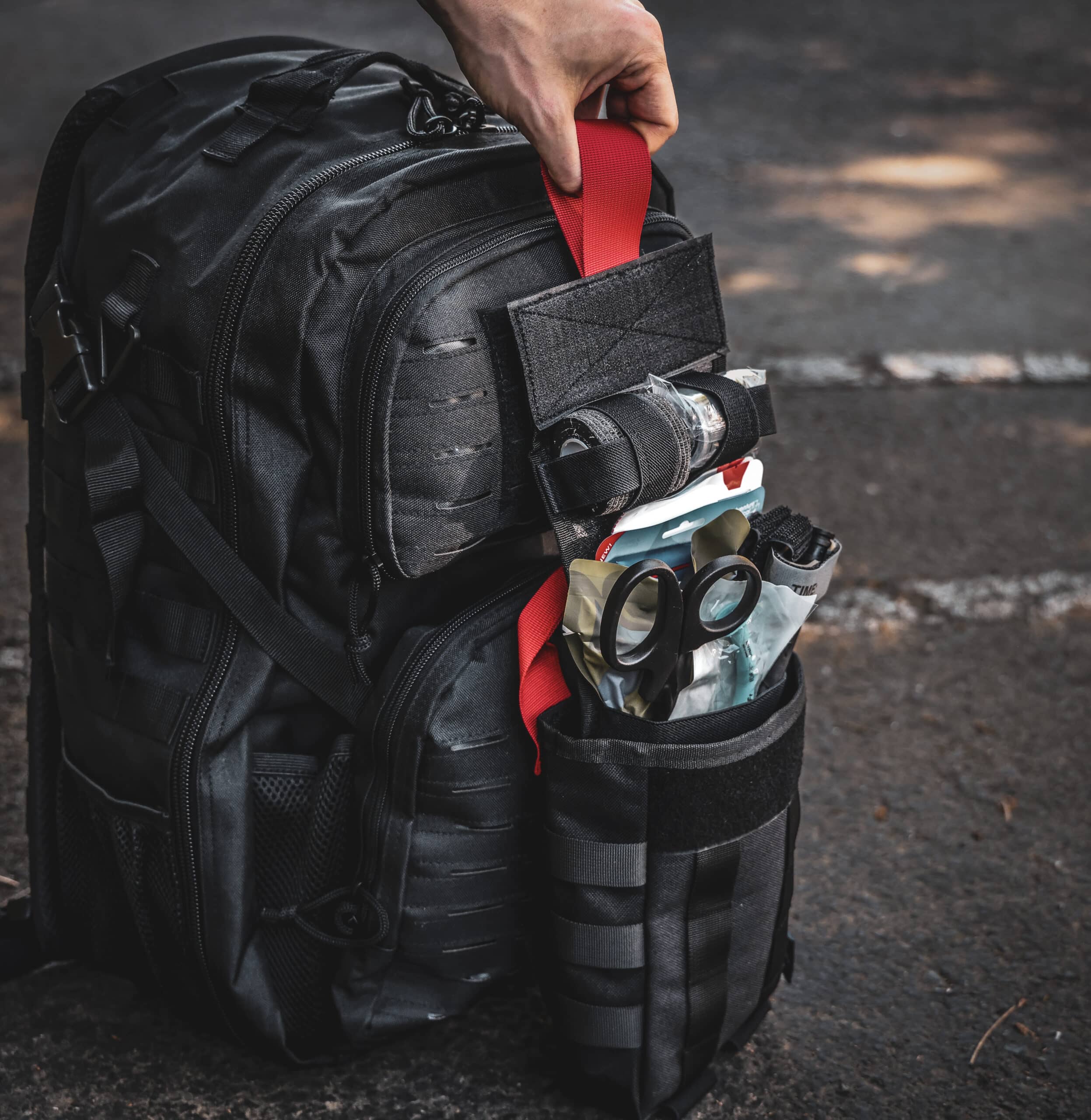 Fully stocked black tactical IFAK pouch attached to MOLLE backpack, displaying trauma shears, QuikClot gauze, and emergency medical gear ready for rapid deployment.