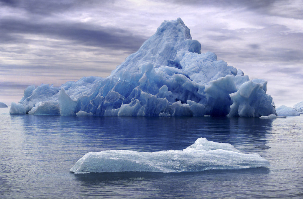 Massive blue iceberg floating in calm, icy waters with a cloudy sky overhead, symbolizing the majesty and isolation of polar landscapes