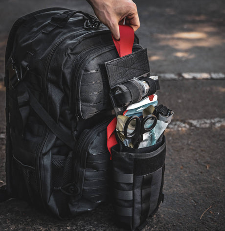 Fully stocked black tactical IFAK pouch attached to MOLLE backpack, displaying trauma shears, QuikClot gauze, and emergency medical gear ready for rapid deployment.